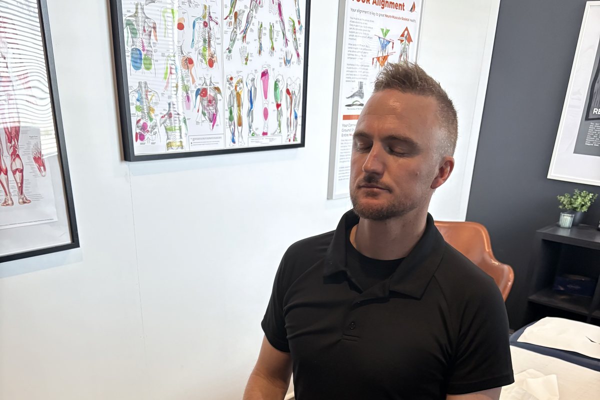 Dale Peirson sitting on the edge of the treatment table with his eyes closed, anatomy charts on the wall behind him